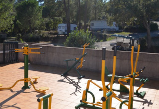 Udendørs fitnessudstyr på en solrig terrasse med træer og campingvogne i baggrunden i en feriepark.