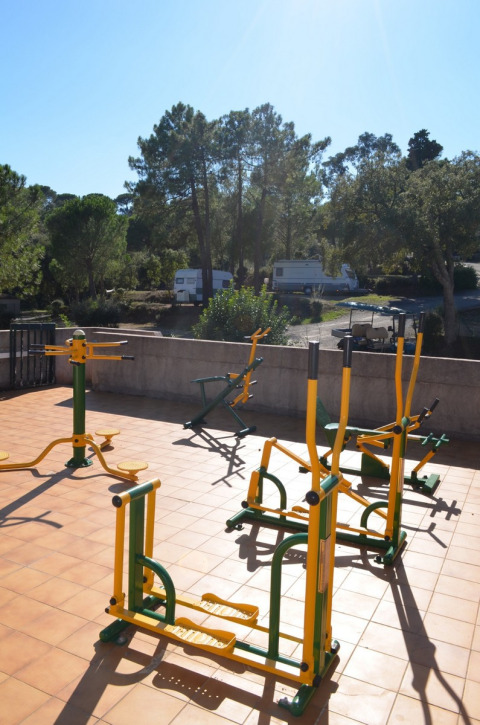 Equipo de gimnasio al aire libre en una terraza soleada, con árboles y autocaravanas al fondo en un parque vacacional.