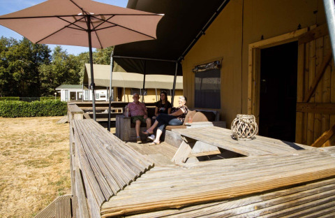 Tres personas descansan en la terraza de una tienda safari en Camping Roland, Países Bajos, bajo una sombrilla.