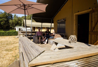 Tre personer slapper af på terrassen ved en safaritelt-lodge på Camping Roland, Holland, under en parasol.