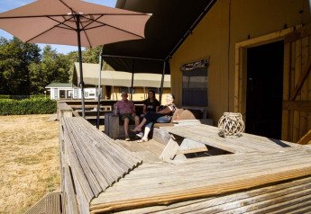 Tre personer slapper af på terrassen ved en safaritelt-lodge på Camping Roland, Holland, under en parasol.