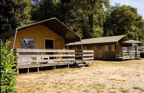 Lodge con tende safari e verande in legno su prato secco, circondato da alberi e vegetazione verde.