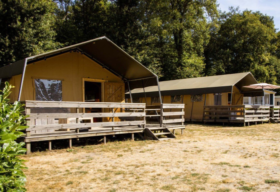 Lodge tente safari avec terrasses en bois sur une pelouse sèche, entourée d’arbres et de verdure.