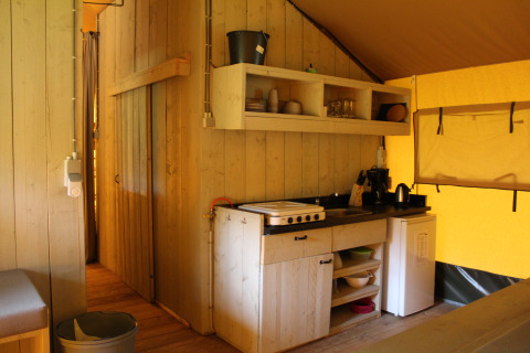 Binnenaanzicht van een safaritent keuken met houten panelen, planken, kookplaat en koelkast in België.