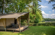 Tienda safari junto a un lago con terraza y sillas en entorno verde en Familiepark Goolderheide, Bélgica.