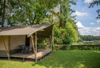 Safari tent by a lake with a deck and chairs in a green setting at Familiepark Goolderheide in Belgium.