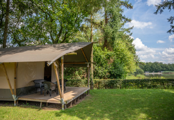 Safari tent by a lake with a deck and chairs in a green setting at Familiepark Goolderheide in Belgium.
