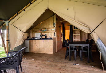 Interno di una tenda safari con cucina in legno, tavolo da pranzo, sedie in plastica e porte aperte.