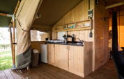 Cuisine de tente safari avec meubles en bois, gazinière, évier, frigo et vue sur la pelouse extérieure.