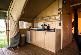 Cuisine de tente safari avec meubles en bois, gazinière, évier, frigo et vue sur la pelouse extérieure.