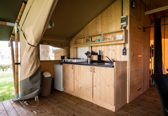 Safari tent keuken met houten kasten, fornuis, koelkast, spoelbak en uitzicht op groene tuin.