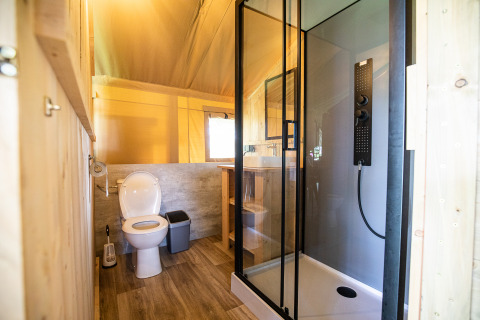 Baño moderno en una tienda tipo safari con ducha, inodoro, cubo de basura, ventana y detalles de madera.