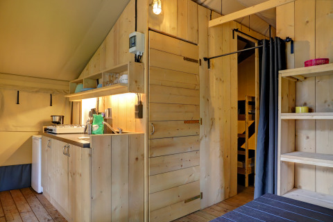 Interno di tenda safari con pareti in legno, piccola cucina e scaffali al Familiepark Goolderheide, Belgio.