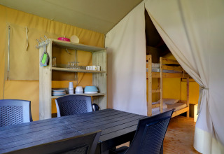 Interior de una tienda safari con mesa de comedor, sillas, estantes y litera en Familiepark Goolderheide, Bélgica.