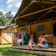 Børn leger på verandaen af et safaritelt med en hund, tøj og træer i baggrunden, Safari tent + sanitet.