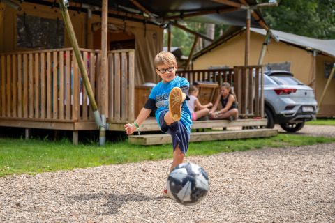 Niño patea una pelota frente a una tienda safari con sanitario en Familiepark Goolderheide, Bélgica.