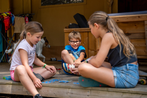 Bambini che giocano a carte sul portico di una tenda safari con servizi igienici a Familiepark Goolderheide.