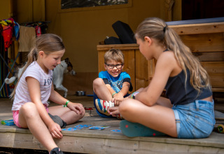 Bambini che giocano a carte sul portico di una tenda safari con servizi igienici a Familiepark Goolderheide.