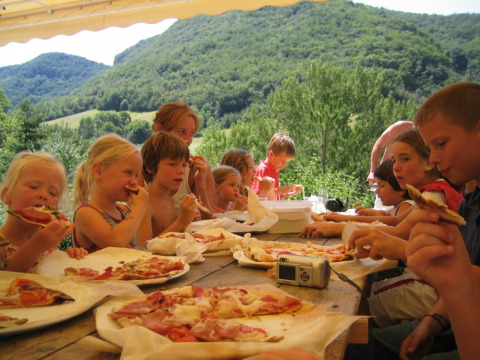 Niños comiendo pizza en una mesa al aire libre con vistas montañosas en Camping Le Gallo Romain, Francia.