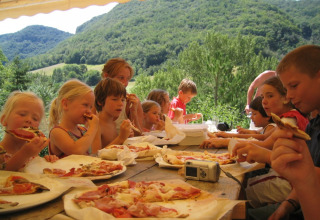 Børn nyder pizza ved et udendørs bord omgivet af grønne bjerge på Camping Le Gallo Romain i Frankrig.