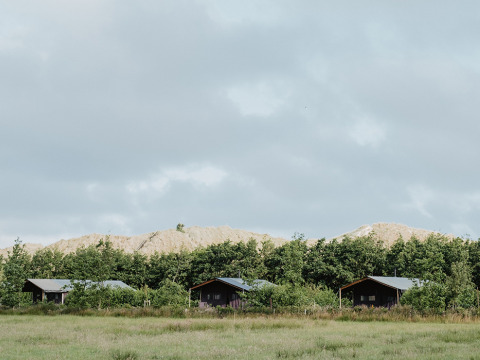 Three wooden cabins sit on a grassy field with trees and sandy hills behind under a cloudy sky.