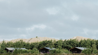 Drie houten huisjes op een grasveld met bomen en duinen op de achtergrond onder een bewolkte lucht.