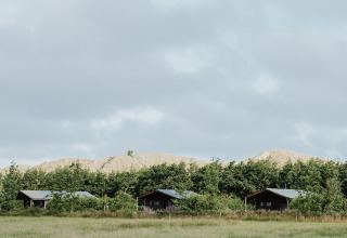 Three wooden cabins sit on a grassy field with trees and sandy hills behind under a cloudy sky.