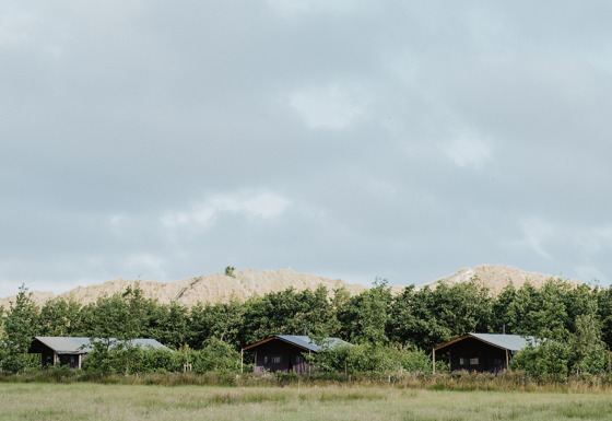 Tre træhytter står på en grøn mark med træer og bakker i baggrunden under en overskyet himmel.