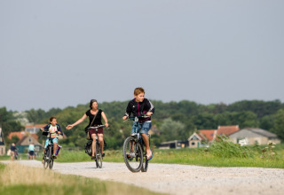Three people are cycling on a gravel path near Oosterend, Friesland, Netherlands, with houses in the background.