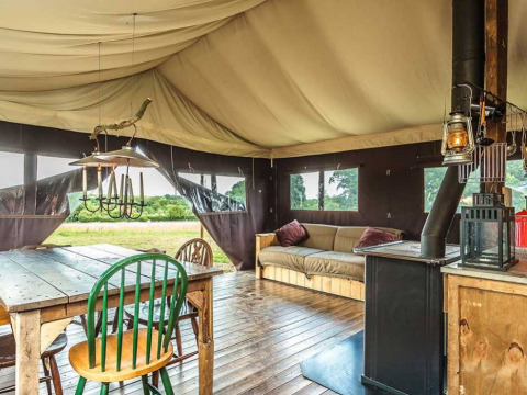 Interior de una tienda glamping con suelo de madera, mesa de comedor, sofá y estufa en un parque vacacional.
