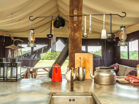 Rustieke glampingkeuken met waterketel, lantaarns en keukengerei, zicht op park via tentramen.