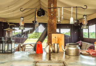 Rustieke glampingkeuken met waterketel, lantaarns en keukengerei, zicht op park via tentramen.