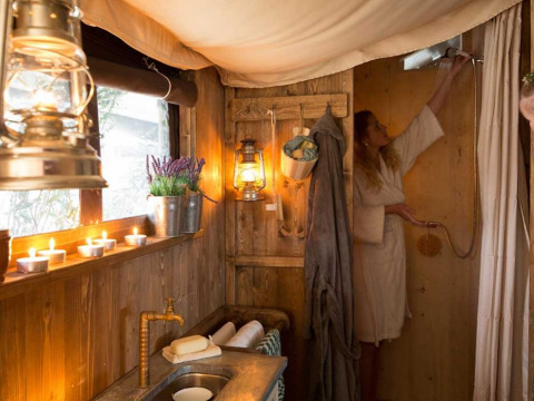 Salle de bain chaleureuse en glamping avec murs en bois, bougies, lanternes et femme en peignoir.