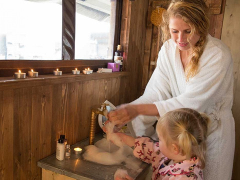 Mutter und Kind im Bademantel waschen Hände an einem Waschbecken im Glamping-Holzhaus mit Kerzen.