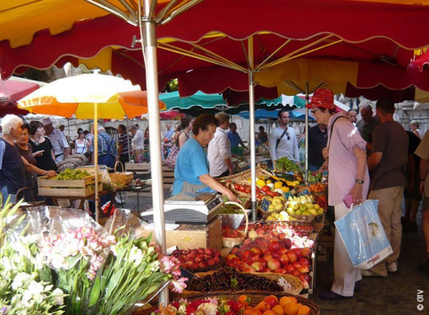 Escena de mercado en Camping lac Aux Oiseaux, Occitania, Francia, con frutas frescas y sombrillas coloridas.