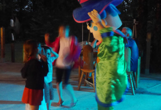 Børn leger og danser med en maskot i farverigt kostume om aftenen på Camping lac Aux Oiseaux i Occitanie.
