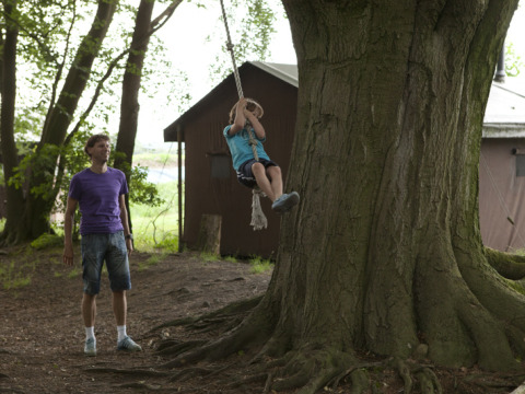 Man watches a child swing on a rope by a large tree at Feather Down Het Wesselink in Salland, Overijssel.