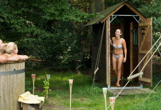 Vrouw in badpak komt uit een houten sauna, twee mensen genieten van een buitenbad in een groene omgeving.