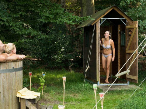 Mujer en traje de baño sale de una sauna de madera, mientras dos personas se relajan en una bañera exterior.