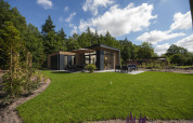 Et moderne lodge med store vinduer, terrasse og havemøbler omgivet af grønne træer og velplejet græsplæne.