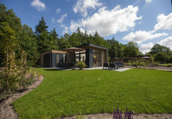 Modern lodgeverblijf met grote ramen, een terras en tuinmeubilair te midden van groen en bomen.