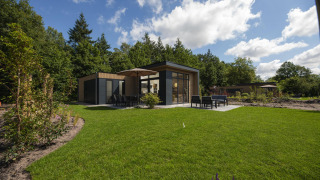 Et moderne lodge med store vinduer, terrasse og havemøbler omgivet af grønne træer og velplejet græsplæne.