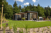 Moderne Lodge mit Terrasse und Gartenmöbeln, umgeben von Rasen und Bäumen bei sonnigem Wetter.