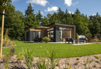 Moderne lodge met grote ramen, terras en tuinmeubilair, omringd door grasveld en bomen.