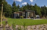 Moderne lodge med store vinduer, terrasse og havemøbler, omkranset af grønt græs og træer.