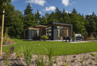 Moderne Lodge mit Terrasse und Gartenmöbeln, umgeben von Rasen und Bäumen bei sonnigem Wetter.