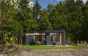 Lodge moderno con ampie vetrate e patio arredato, immerso nel verde tra alberi in una giornata soleggiata.