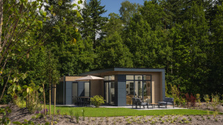 Moderne lodge med store vinduer, terrasse og havemøbler, omgivet af grønne træer og natur under blå himmel.