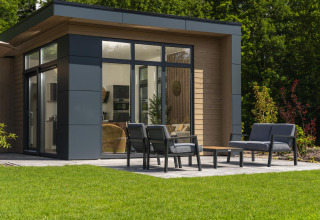 Lodge moderne avec grandes fenêtres, terrasse aménagée, mobilier d’extérieur et pelouse soignée devant.