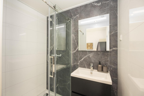 Baño moderno en una posada con ducha de vidrio, lavabo oscuro, gran espejo y azulejos efecto mármol.